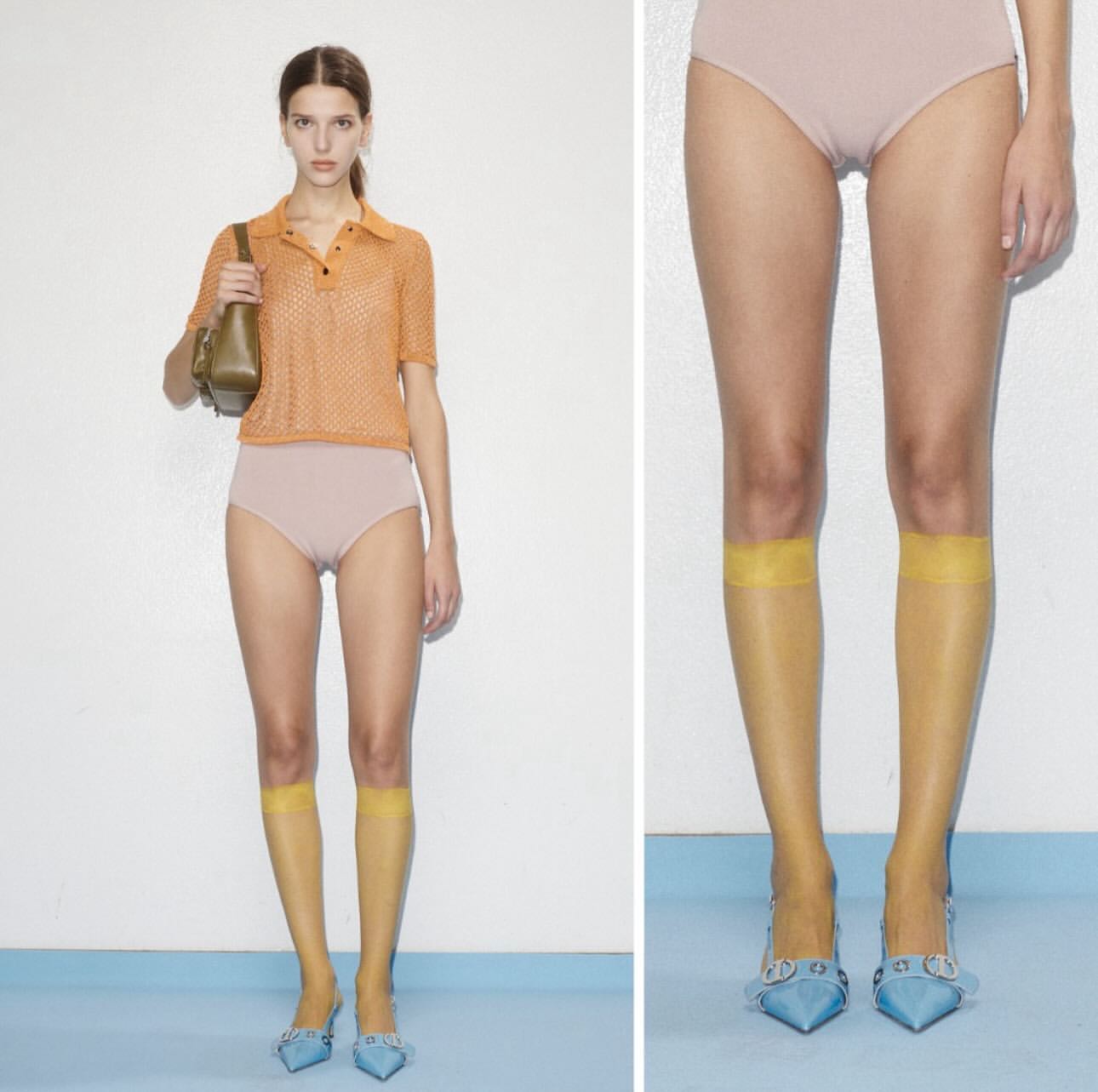 A young, light-skinned female fashion model poses in a studio with an orange top, light-pink bottoms, and yellow hosiery with matching light-blue heels.