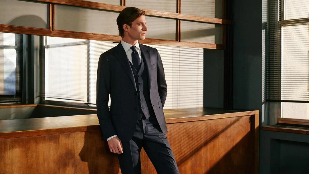 A confident man in a three-piece suit poses indoors near a window, showcasing elegant menswear fashion in a well-lit setting.