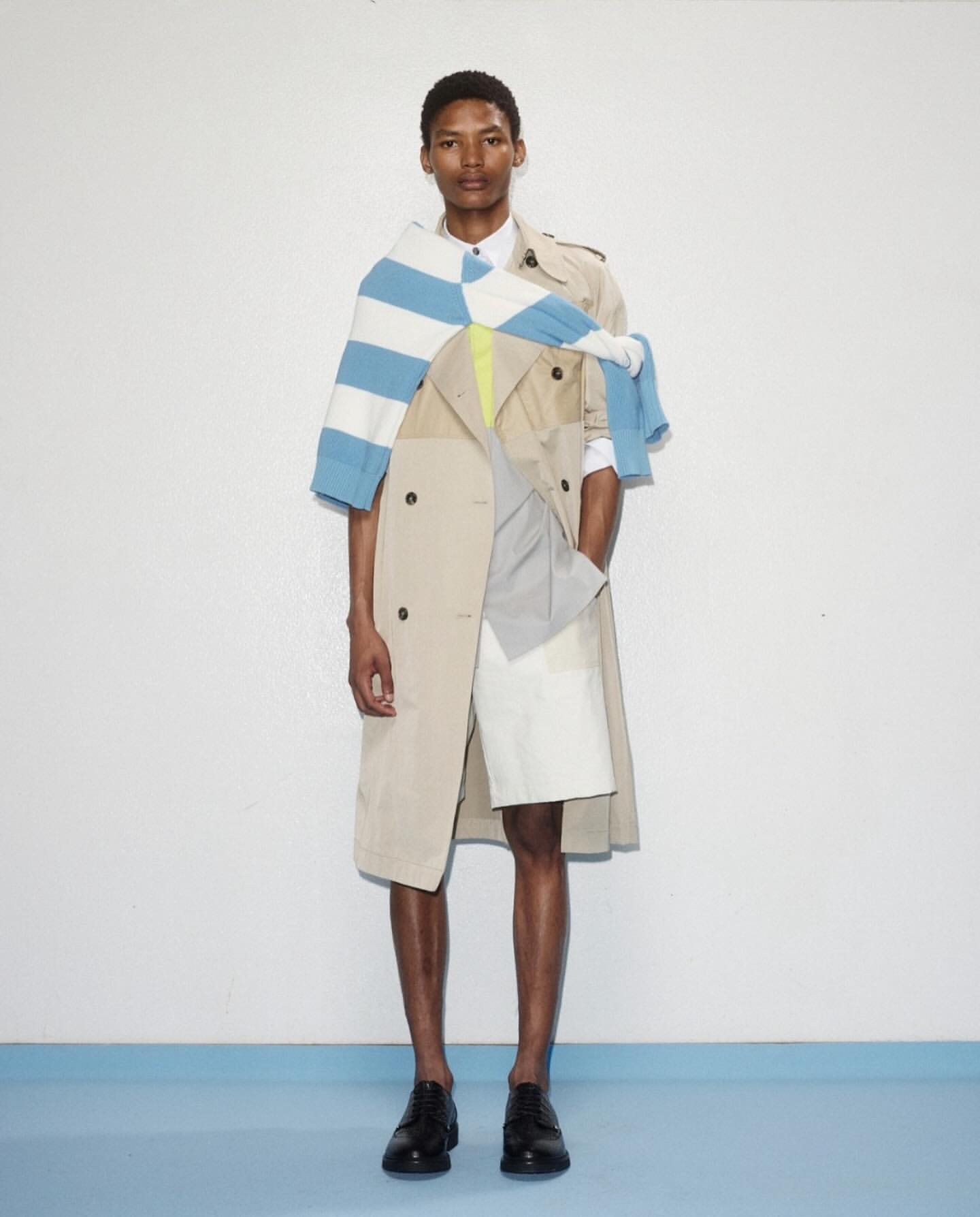 Full shot of a slender man in a beige trench coat, white shirt, blue striped sweater, and black shoes against a white and blue backdrop.