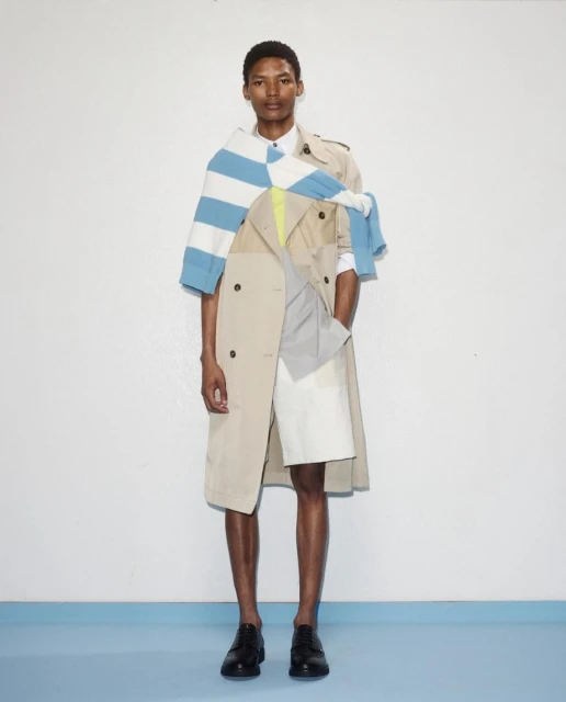 Full shot of a slender man in a beige trench coat, white shirt, blue striped sweater, and black shoes against a white and blue backdrop.