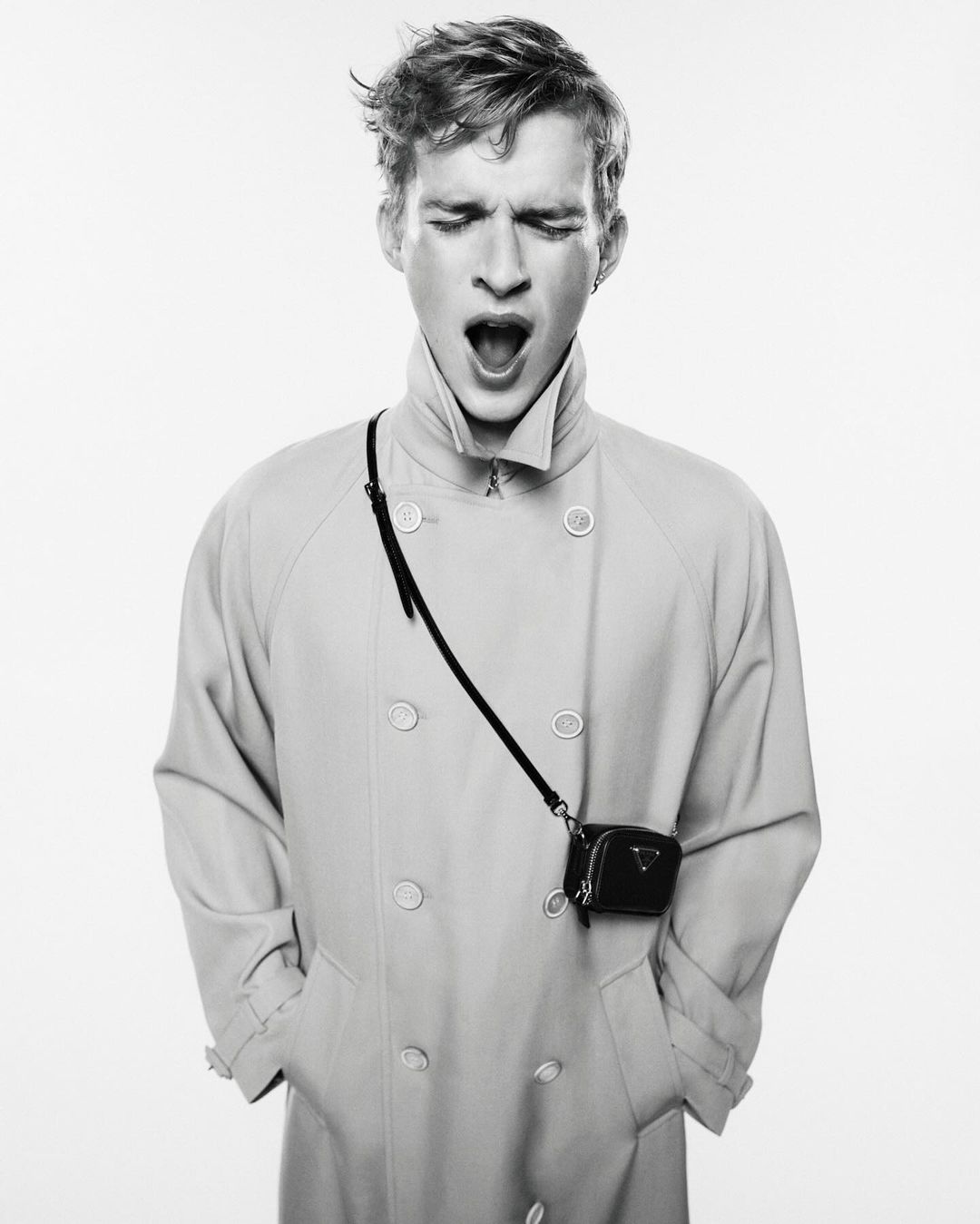 Black and white fashion portrait of a young adult male model yawning while wearing a light-colored trench coat and a small purse.
