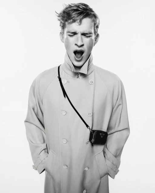 Black and white fashion portrait of a young adult male model yawning while wearing a light-colored trench coat and a small purse.
