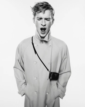 Black and white fashion portrait of a young adult male model yawning while wearing a light-colored trench coat and a small purse.