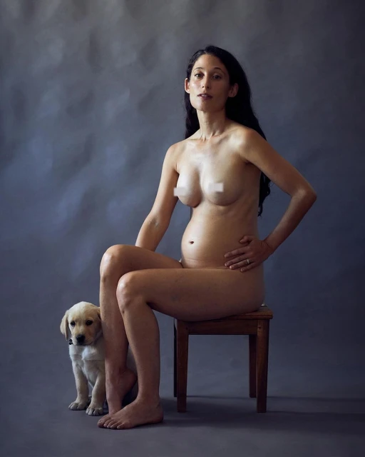 Nude pregnant woman sits on wooden chair indoors with a yellow Labrador puppy at her feet against a mottled gray background.