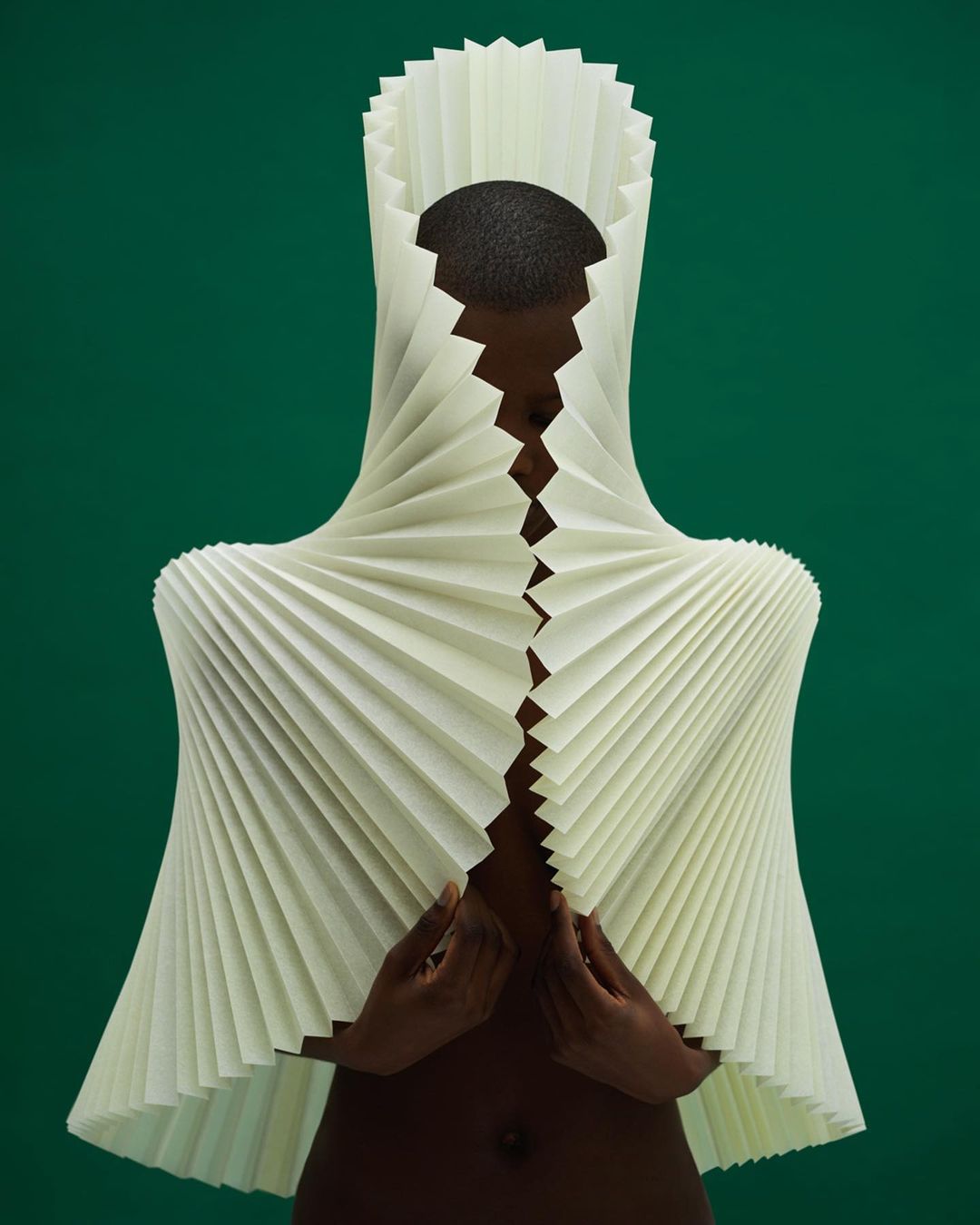 Model showcasing an avant-garde paper dress with pleated texture and unique headpiece, set against a solid green background.