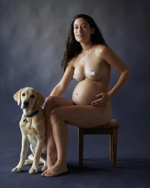 Pregnant woman sits next to her dog in a studio. She is nude and smiling with dark, wavy hair. Her yellow lab sits attentively.