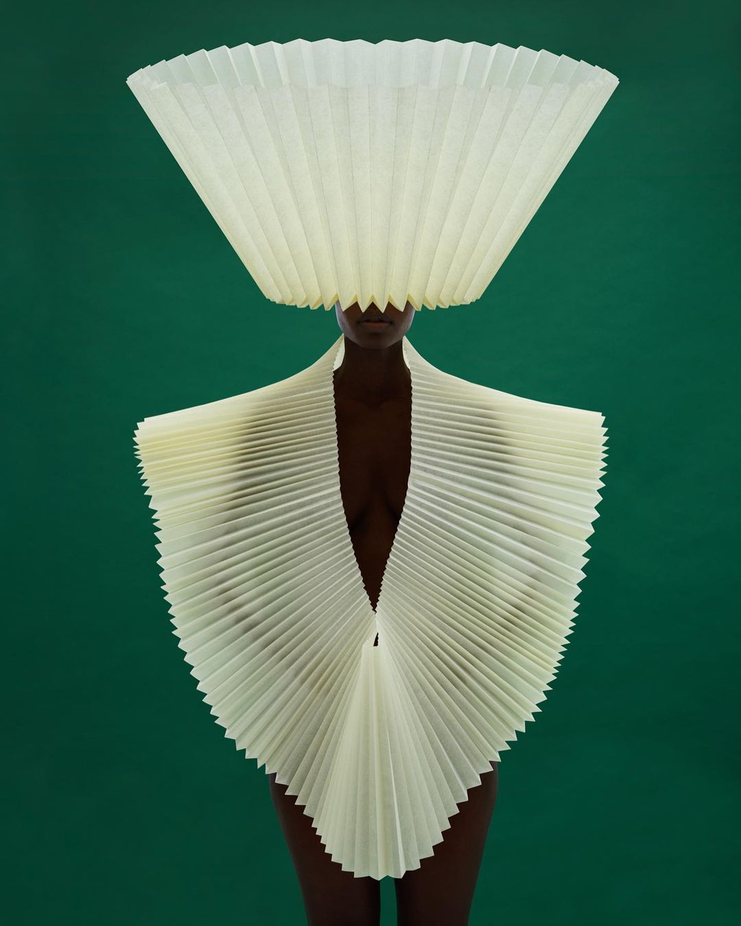 Stunning fashion shot: an adult female in an avant-garde, pleated paper dress and headpiece against a vibrant green backdrop, artistic expression.