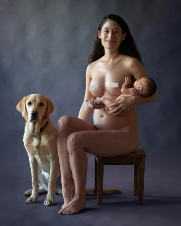Woman sits on wooden stool, holding infant, and smiling beside a golden retriever dog against a mottled gray background in a studio.