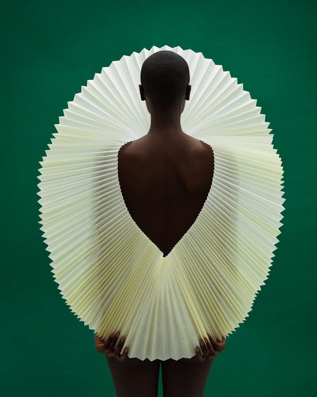 Fashionable African woman in dramatic pleated circular collar, shot from behind against a deep green backdrop in a studio setting.