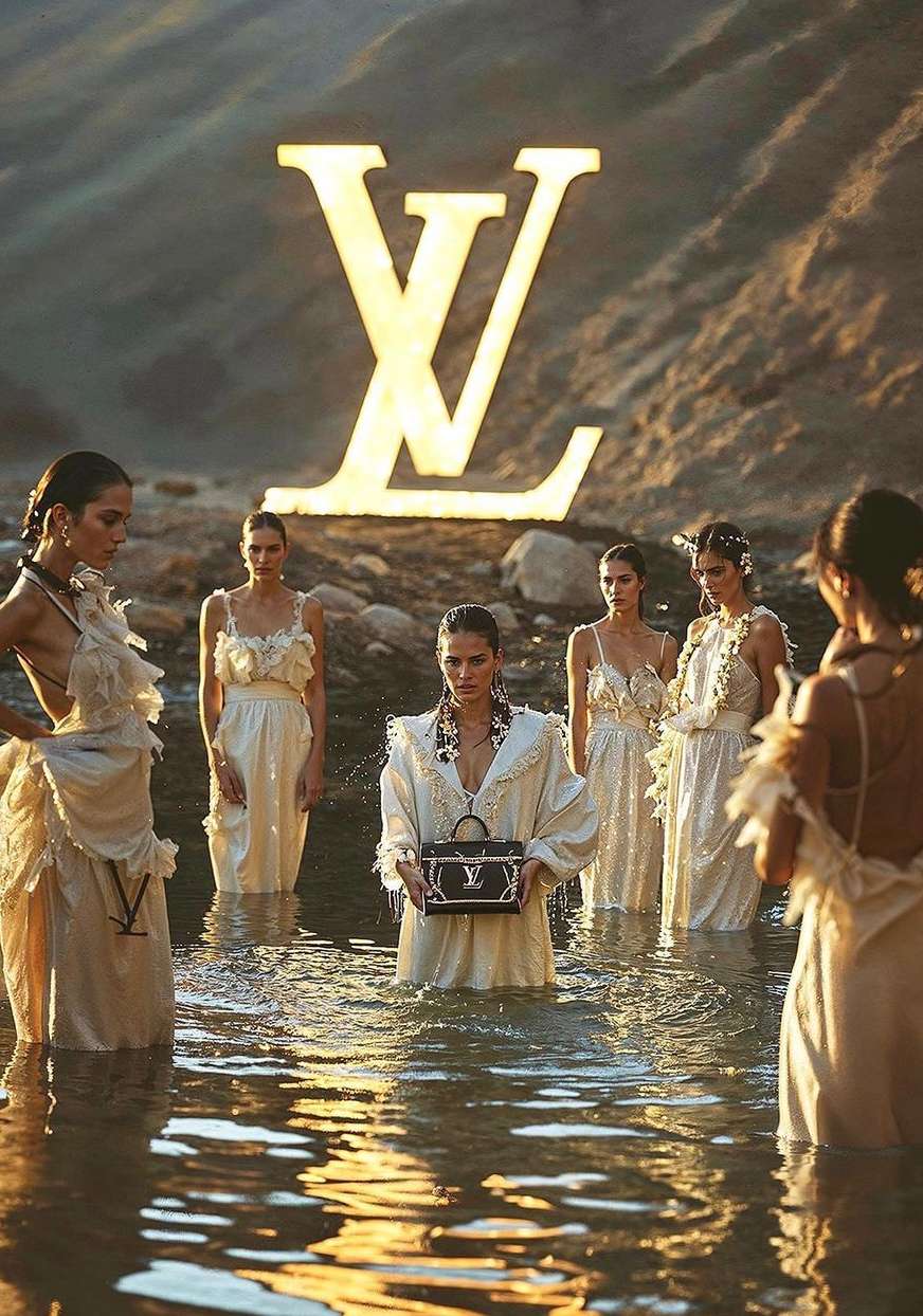 Six women stand in water wearing light-colored dresses, holding a handbag in front, a large Louis Vuitton logo in the background.