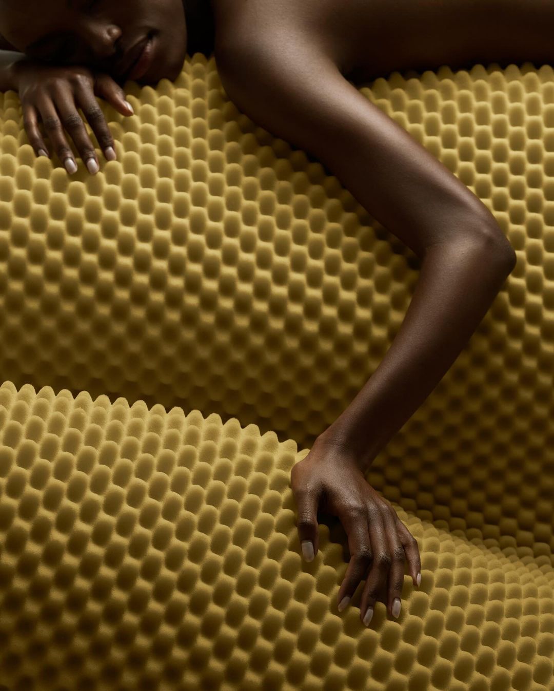 Close-up of a Black woman lying on a yellow textured surface, showcasing her arm, hand, and face in a studio-like setting. 