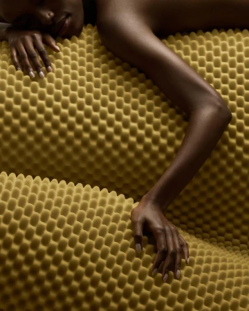 Close-up of a Black woman lying on a yellow textured surface, showcasing her arm, hand, and face in a studio-like setting. 