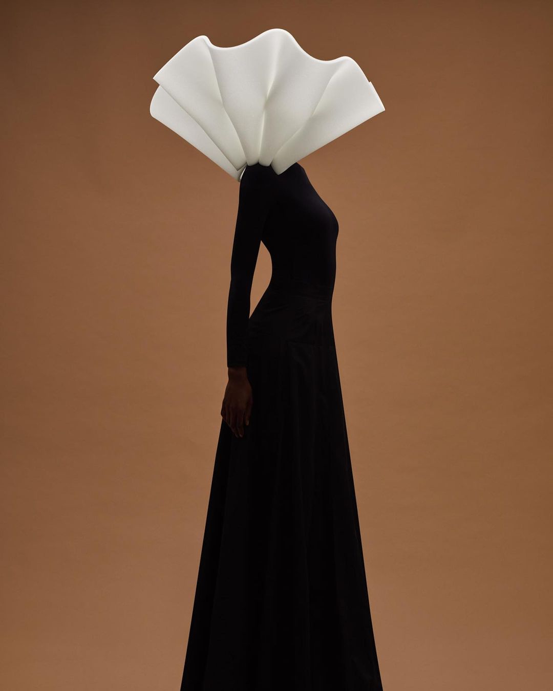 Woman in black dress with a sculptural white headpiece against a tan background in a fashion editorial style with dramatic lighting.