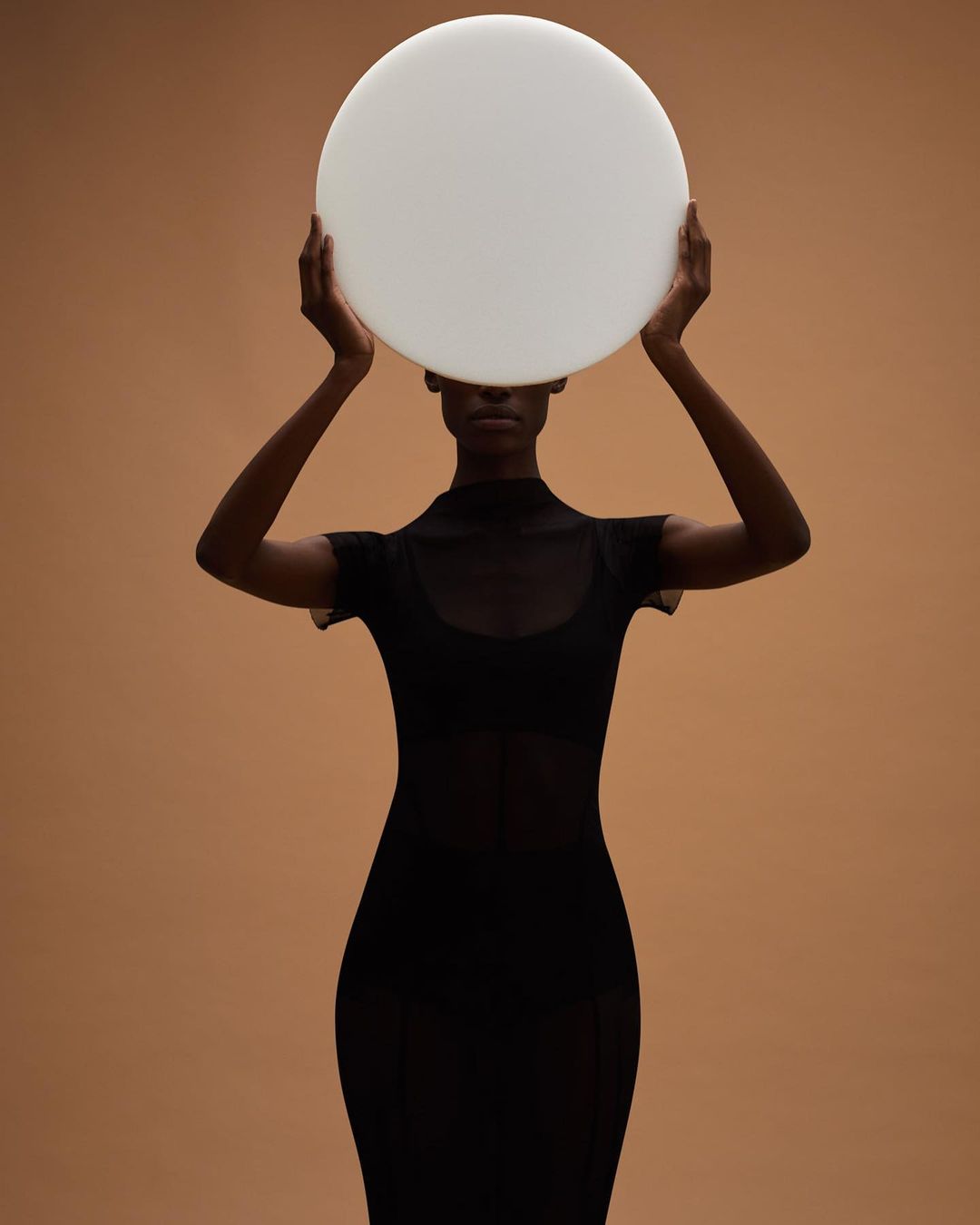 An elegant Black woman in a sheer dress holds a white circular disc over her head against a neutral tan background in a studio.