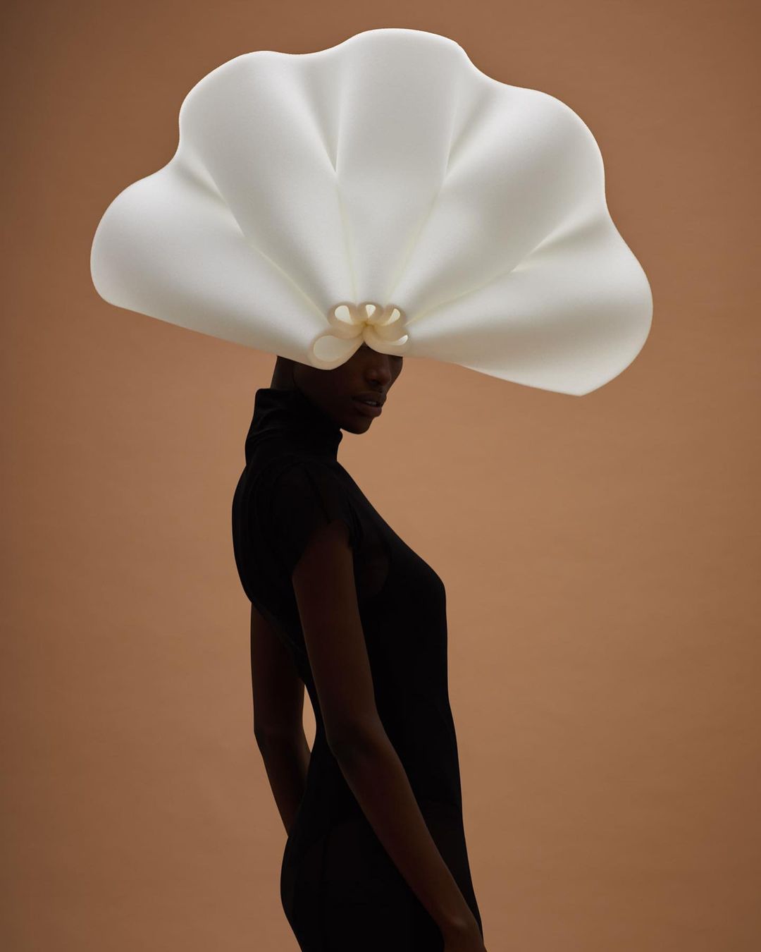 Woman in a black dress poses in front of a tan background wearing a large, white, sculptural hat in a fashion forward style.