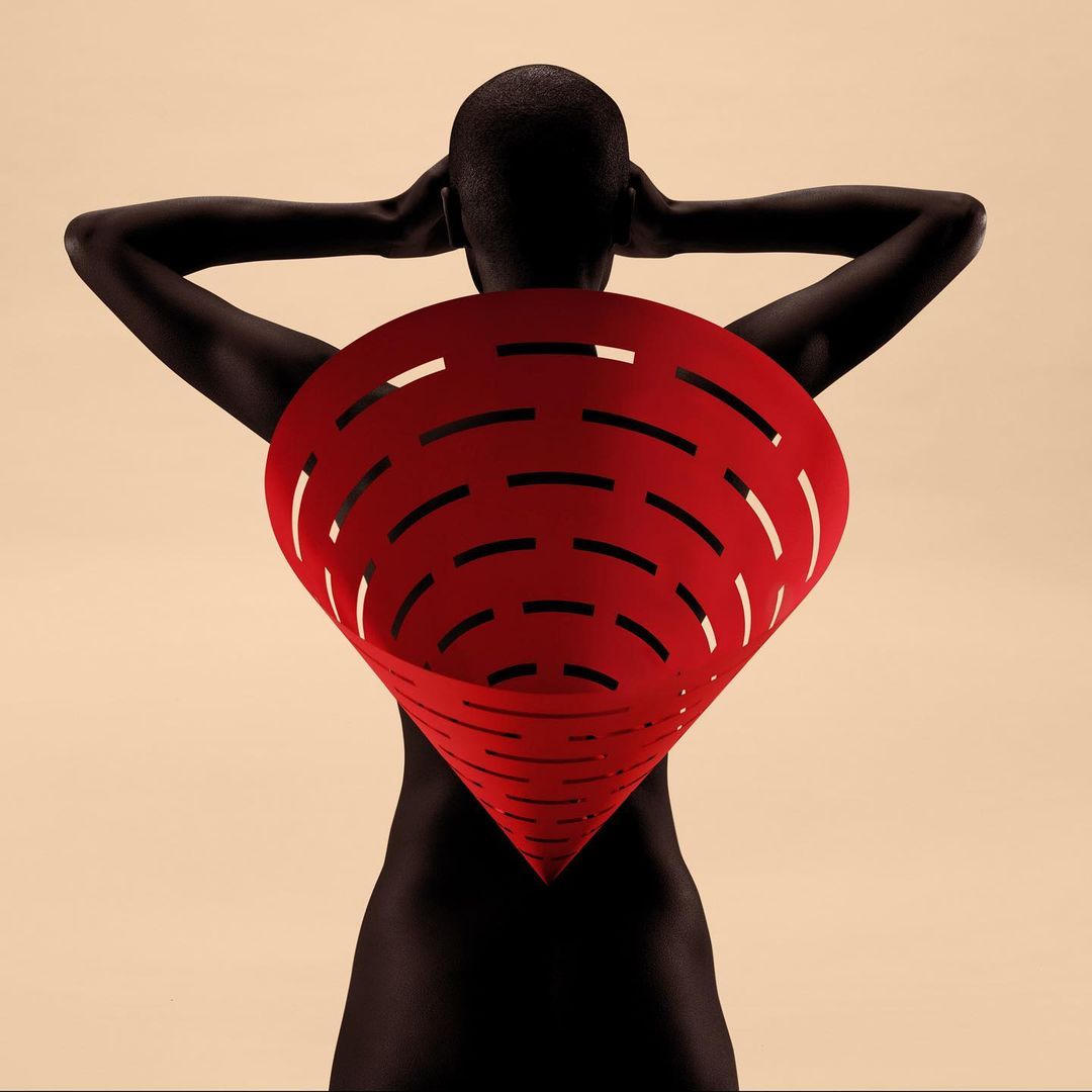 Conceptual studio shot of an adult female model with dark skin, facing away, wearing a large red paper cone from her upper back.