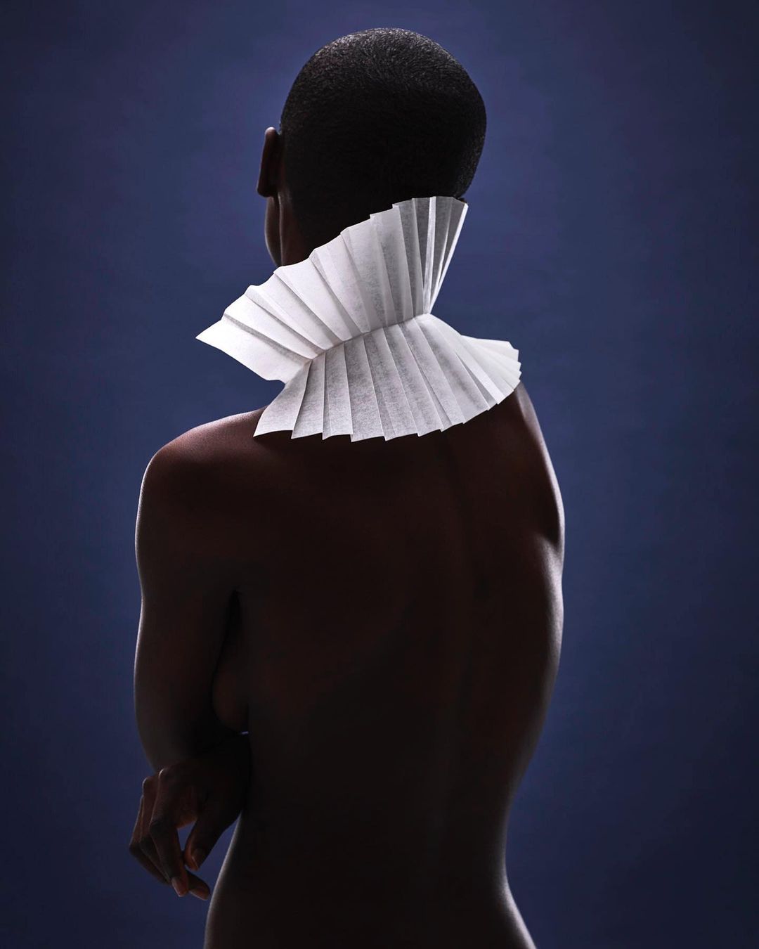Striking portrait of a shaved-head person wearing a decorative white paper ruff collar against a deep blue background in a studio setting.