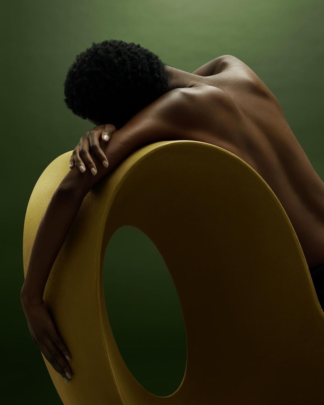 Back view of a Black woman with short hair resting on a mustard-yellow modern chair against a dark green background in a studio setting.