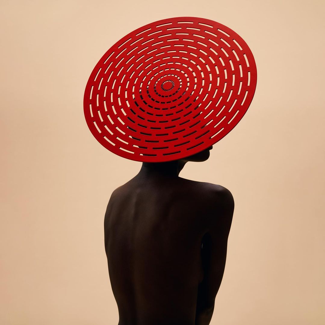 Back view of a dark-skinned woman wearing a large red circular hat with concentric circle cutouts against a light beige background.