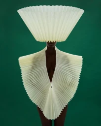 Model wearing a large pleated lampshade-style headpiece and sculptural white pleated collar against a solid dark green background.