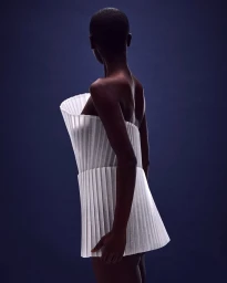 Model with a short-cropped hairstyle wearing a structured white pleated top against a dark blue background