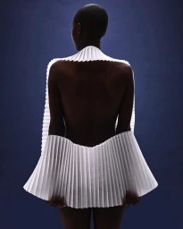 Model seen from behind wearing an architectural white pleated paper collar against a dark blue background