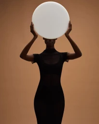 Model holding a large white circular disc over her head, wearing a black sheer dress against a warm brown background.