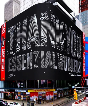 Outdoor view of a large-scale black and white mural in Times Square that thanks essential workers above the storefronts on a city street.