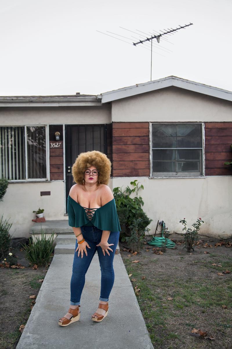 Portrait of a woman with an afro and glasses, wearing a green top and jeans, posing on a walkway in front of a house.