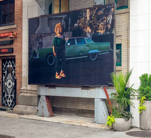 Outdoor billboard featuring a photograph of an adult female in a green outfit and orange shoes standing next to a green vintage car.