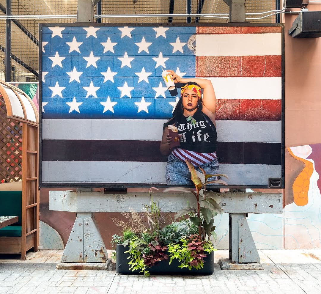 Mural of a woman holding food and drink in front of a painted American flag with a plant box at the base of the mural.