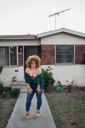 Woman with a voluminous blonde afro wearing a green off-the-shoulder top and blue jeans on a concrete walkway in front of a house.