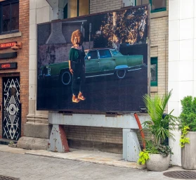 Public art billboard installation featuring a photograph of a woman with an afro posed in front of a vintage green car on a city street.