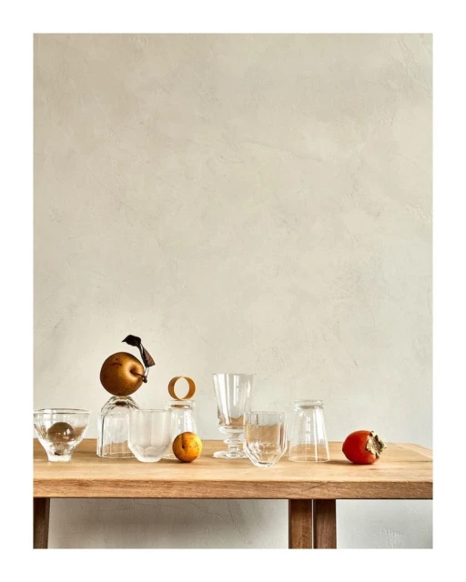 Still life photography showcases clear glasses, a pear with ribbon, and an orange on a wooden table against a neutral background.