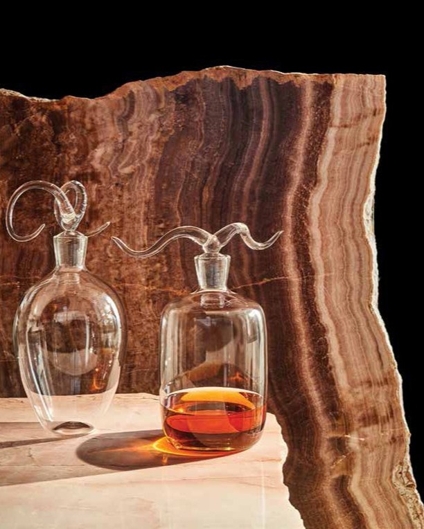 Two glass decanters with stoppers sit on a marble surface against a background of layered brown onyx stone and a black backdrop.