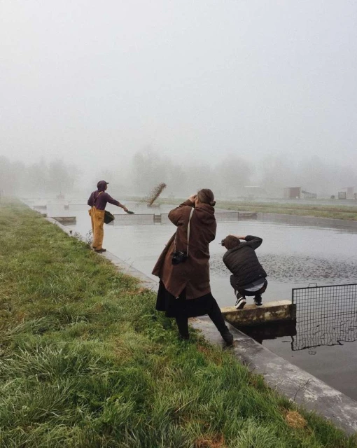 Three people near a pond on a foggy day, one adult male in a hat spreading feed, one adult female taking photo, and another adult female crouched down.