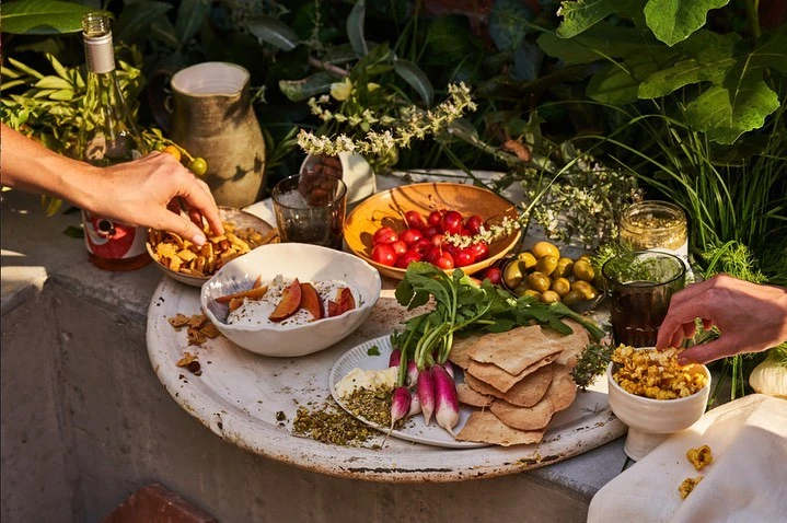 Overhead shot of a white table with bowls and plates of fresh food, including chips, tomatoes, olives, radishes, and a pitcher, outdoors.