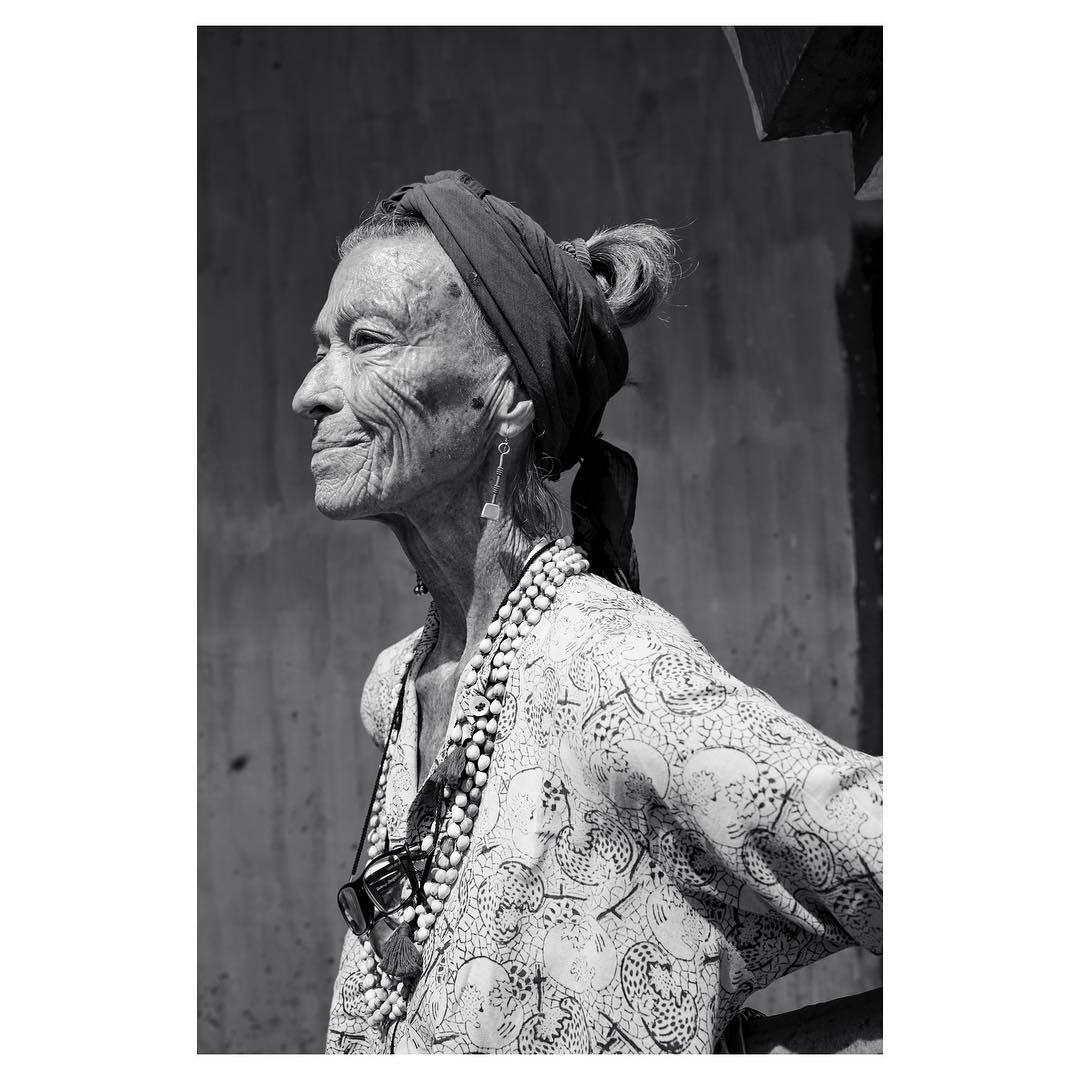 Striking black and white portrait of an elderly woman with a headscarf, bead necklaces, and a patterned top smiling outdoors.
