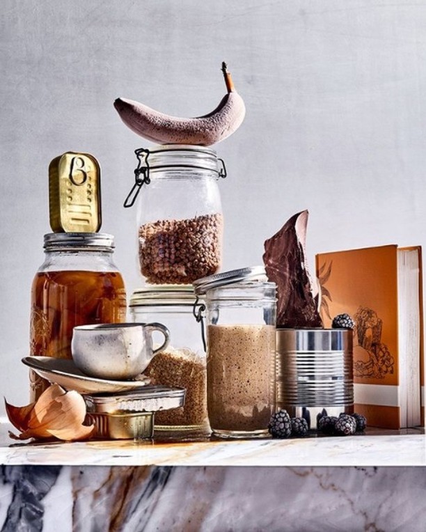 Still life shot featuring a banana, glass jars, canned goods, and a cup, stacked artfully against a light gray background.