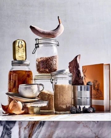 Still life shot featuring a banana, glass jars, canned goods, and a cup, stacked artfully against a light gray background.