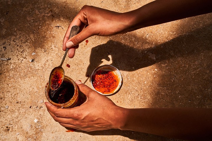 Close-up of hands holding a jar of chili oil with a spoon scooping into another open jar, shot on a tan textured surface.
