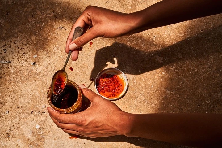 Close-up of hands holding a jar of chili oil with a spoon scooping into another open jar, shot on a tan textured surface.