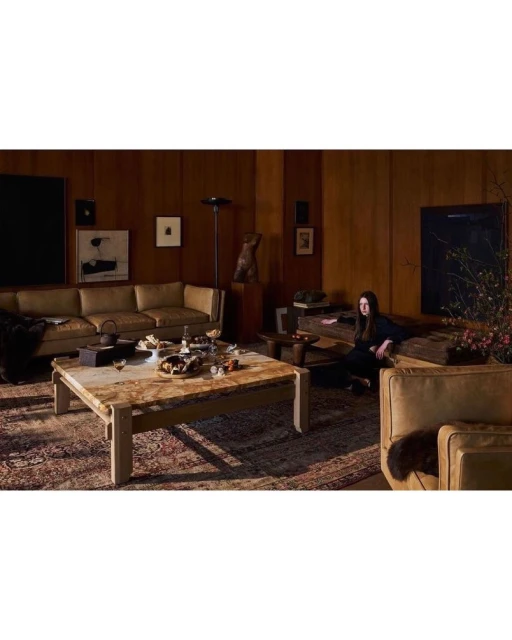 Interior of a home with an adult female sitting near a tan couch and coffee table displaying food, wood paneling on the walls.