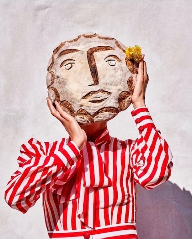 Woman holding decorative bread with facial features, wearing a red and white striped top, against a bright white background.