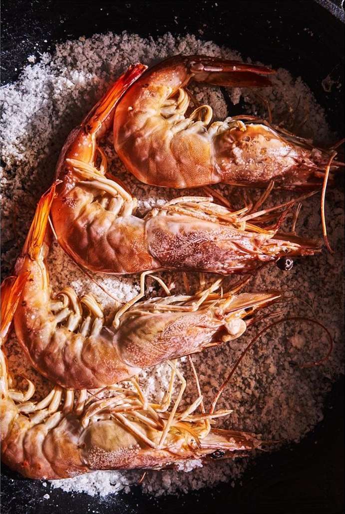 Overhead shot of four cooked shrimp in a bed of salt, filling the dark frame, focusing on culinary presentation and seafood.