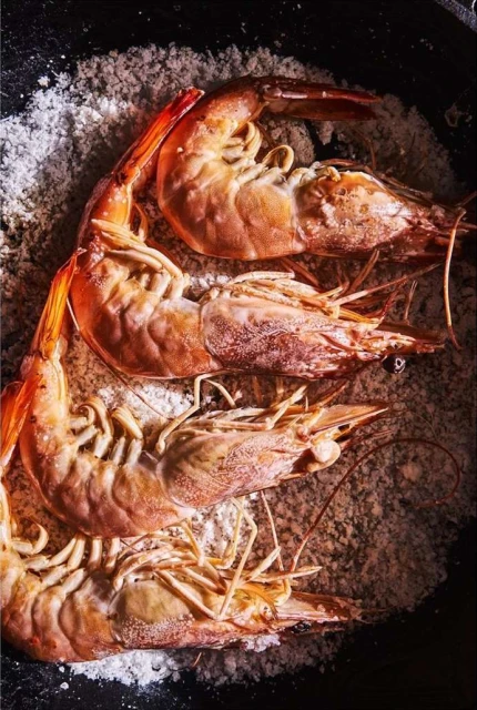 Overhead shot of four cooked shrimp in a bed of salt, filling the dark frame, focusing on culinary presentation and seafood.