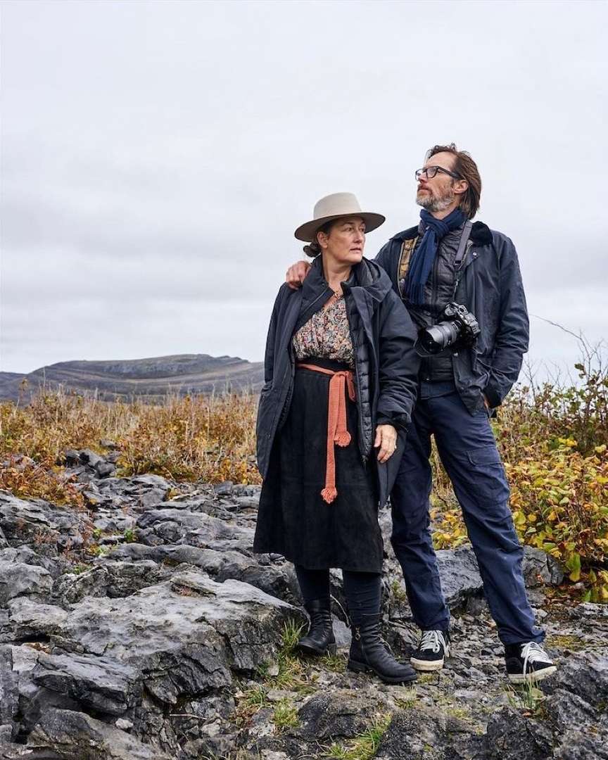 Outdoor portrait of a couple standing on a rocky terrain with mountains in the background, wearing jackets and hats, looking off into the distance.