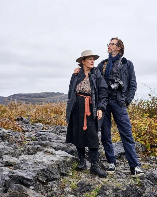 Outdoor portrait of a couple standing on a rocky terrain with mountains in the background, wearing jackets and hats, looking off into the distance.
