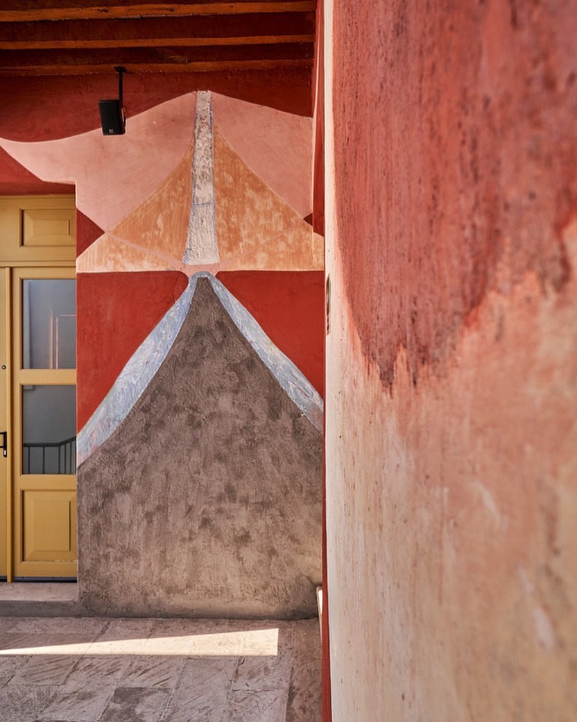 Exterior shot of a terracotta building with an abstract geometric design in shades of red, brown, and beige next to a mustard-yellow door.