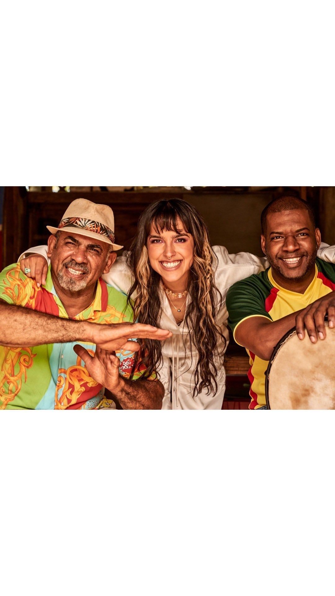 Three musicians, two men and one woman, pose together indoors. The men hold instruments and the woman wears a white outfit with jewelry.